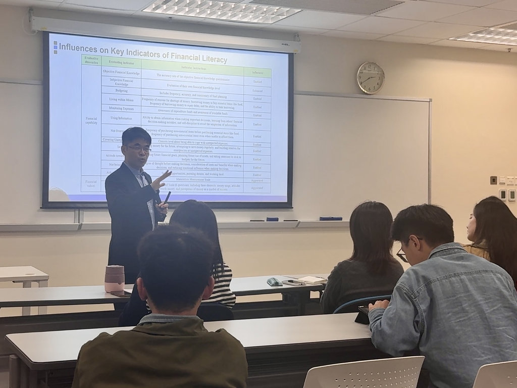 Lecture scene &mdash; a speaker presenting to students with a slide titled 'Influences on Key Indicators of Financial Literacy'. Note: AI-generated content may be incorrect. / 注意：AI 生成的内容可能不正确。